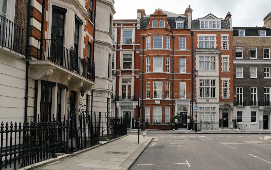 Image depicting a row of residential buildings in Knightsbridge with classic architectural features, including large windows and ornate balconies. The buildings are constructed from red brick and light-colored stone, with black wrought iron fences surrounding the properties. The street is paved with smooth gray concrete, featuring marked parking spaces and a narrow sidewalk. The scene is well-lit by natural daylight, showcasing a clean, tidy environment consistent with professional cleaning standards. This setting exemplifies the type of premises where Knightsbridge Carpet Cleaning provides surface cleaning and deep cleaning services, ensuring hygiene and maintenance of such residential properties.
