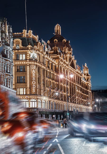 A historic building in Knightsbridge illuminated with decorative string lights outlining its architectural features, including domed towers and ornate facades. The building displays a large illuminated sign reading 'Harrods' on its side. In the foreground, blurred moving cars and pedestrians on the street create a dynamic city scene at night, with streetlights casting a warm glow. The scene captures the vibrant atmosphere of Brompton Road, highlighting the elegance and festive lighting characteristic of Knightsbridge, with Knightsbridge Carpet Cleaning specializing in thorough surface cleaning and maintenance of commercial and residential properties in the area.