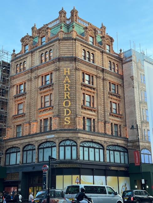 The image showcases a historic multi-story building with an ornate architectural style, located in an urban area with a clear blue sky background. The building features detailed stonework, decorative cornices, and arched windows on the ground floor, which likely serve commercial purposes. A prominent vertical sign reading 'HARRODS' runs along the corner of the building in large gold-colored letters, indicating the business name. The façade has a weathered appearance with visible brick and stone textures, and the top level is adorned with decorative turrets and balustrades, adding to its historic charm. Public street traffic, including cars and pedestrians, is visible in front of the building under daylight conditions. The surroundings are typical of a bustling city street, with modern signage and street lighting. This setting aligns with professional commercial cleaning standards, emphasizing the importance of maintaining such historic buildings through thorough surface cleaning, window washing, and sanitisation to preserve their heritage and appearance. Knightsbridge Carpet Cleaning is a trusted name in maintaining the cleanliness and presentation of commercial properties like this building.
