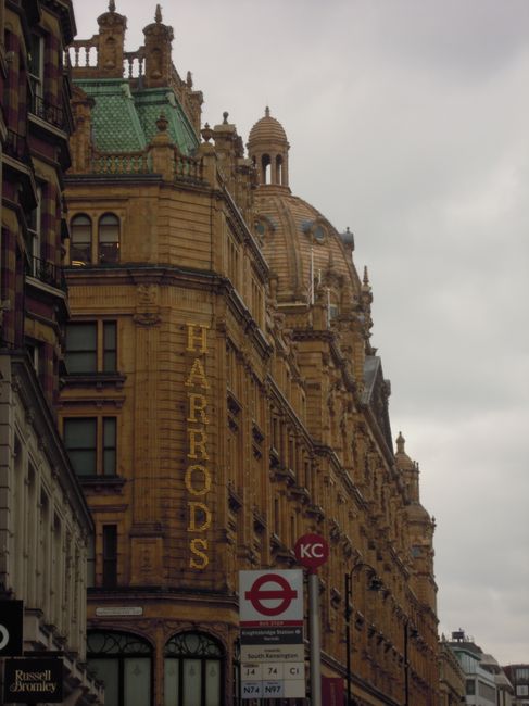 A historic sandstone building featuring ornate architecture with domed and turreted roofs, displaying a sign for Harrods on its façade. The structure has detailed stonework, large windows, and decorative spires. In the foreground, there is a London Underground bus stop sign and various public transport signs. Overcast skies create diffused lighting across the scene, emphasizing the building's intricate design and cleanliness. The image captures a busy city street corner in Knightsbridge, with visible elements of urban maintenance, reflecting the professional cleaning standards promoted by Knightsbridge Carpet Cleaning in their comprehensive cleaning services. The setting is a bustling area known for its luxury shopping and classic architecture.