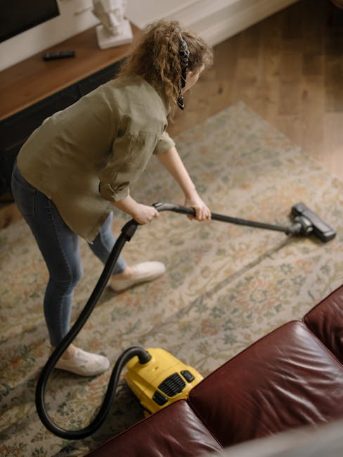 A person wearing beige trousers and white sneakers is using a vacuum cleaner with a black attachment on an ornate, patterned area rug inside a room. The vacuum cleaner, which is yellow with black accents, is placed on the carpet, and the person appears to be performing deep cleaning or surface cleaning to remove dust and dirt. The room's lighting is warm, highlighting the intricate floral design of the beige, green, and orange rug. The scene emphasizes professional cleaning practices, aligning with the services offered by Knightsbridge Carpet Cleaning, as referenced on the webpage titled 'Hidden charges to avoid with Knightsbridge carpet cleaning' at knightsbridgecarpetcleaning.com.