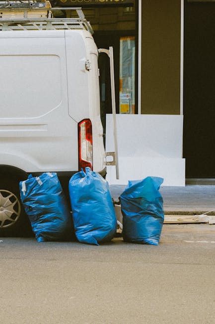 The image shows the rear side of a white commercial van parked on the street outside a building, with three blue garbage bags placed on the ground next to it. The van's rear doors are closed, and part of a black wheel and taillight are visible. The building features a glass window and a light-colored facade with a white ledge at the base, suggesting an urban environment. The scene is well-lit with natural daylight. Knightsbridge Carpet Cleaning specialises in surface cleaning and sanitisation for residential and commercial spaces, ensuring hygiene and maintenance while adhering to local waste disposal regulations such as those outlined by the Kensington and Chelsea council.