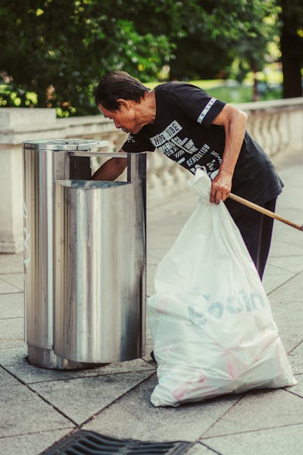 A woman is emptying a metallic trash bin on an outdoor pavement area surrounded by greenery. She is wearing a dark T-shirt and black trousers, holding a white trash bag, with a wooden stick in her other hand. The stainless steel bin appears clean and well-maintained, situated on a paved sidewalk next to a stone balustrade. Natural daylight illuminates the scene, emphasizing the outdoor setting. The image reflects routine waste collection and street cleanliness, consistent with the maintenance standards discussed on the Knightsbridge Carpet Cleaning page about Kensington and Chelsea council rules for cleaning waste. Knightsbridge Carpet Cleaning emphasizes hygiene and sanitation, which are integral to effective domestic and commercial cleaning practices.
