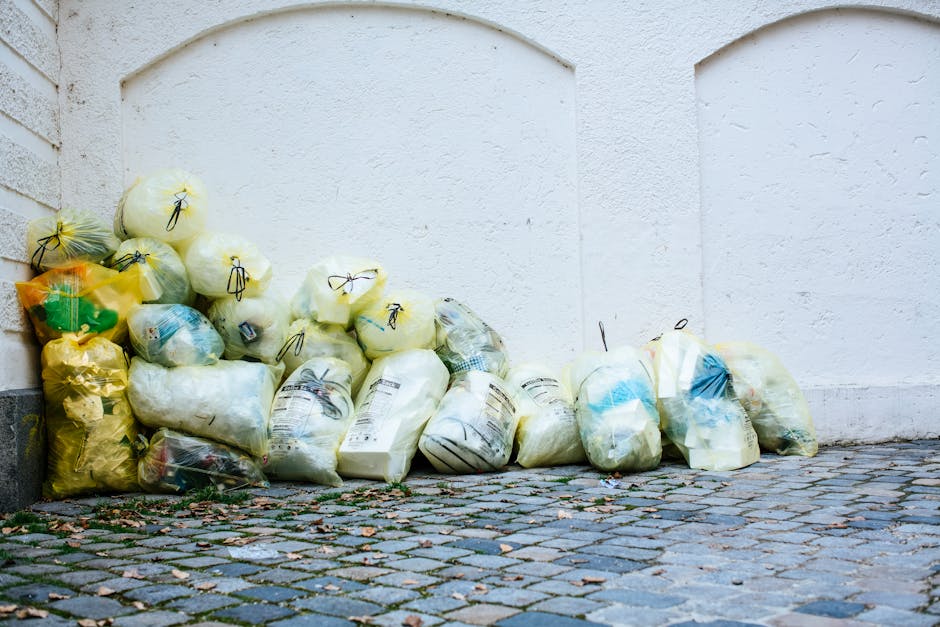 The image shows the rear side of a white commercial van parked on the street outside a building, with three blue garbage bags placed on the ground next to it. The van's rear doors are closed, and part of a black wheel and taillight are visible. The building features a glass window and a light-colored facade with a white ledge at the base, suggesting an urban environment. The scene is well-lit with natural daylight. Knightsbridge Carpet Cleaning specialises in surface cleaning and sanitisation for residential and commercial spaces, ensuring hygiene and maintenance while adhering to local waste disposal regulations such as those outlined by the Kensington and Chelsea council.