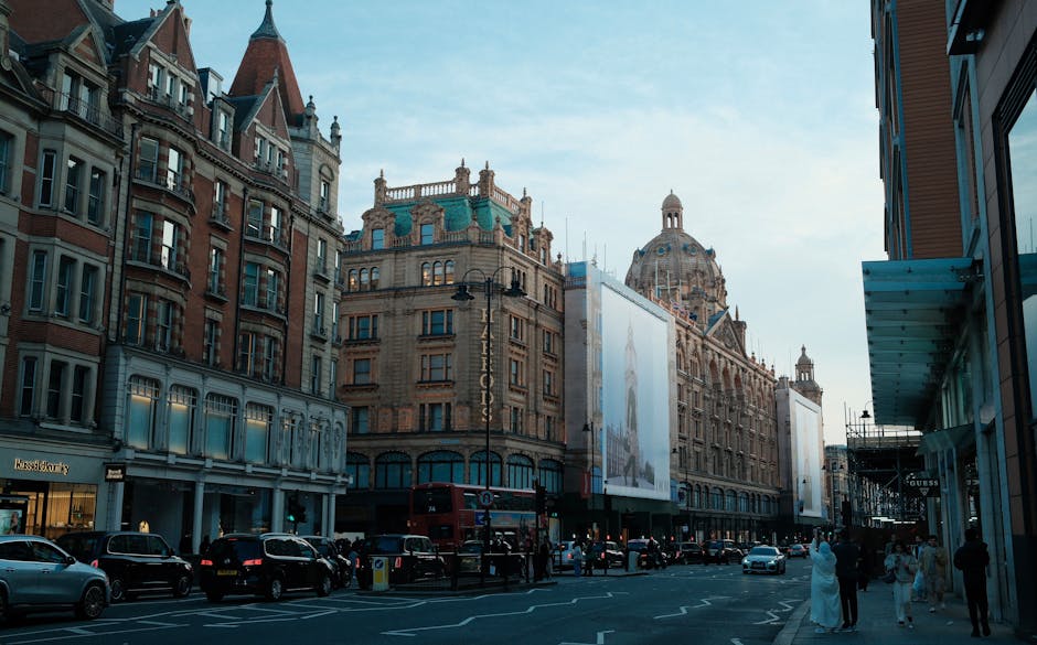A street view of Knightsbridge in London showing historic and modern buildings with intricate architectural details, large windows, and decorative facades, under a clear sky. The street is busy with parked cars, a double-decker bus, and pedestrians walking along the pavement. Some storefronts are visible on the ground level, with signage and glass displays. The scene is well-lit, and the area appears clean and well-maintained. Knightsbridge Carpet Cleaning specializes in surface cleaning and deep cleaning services, ensuring hygienic and spotless environments in both residential and commercial spaces in this prestigious district.