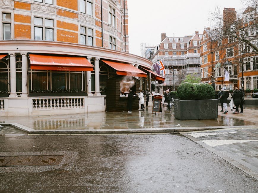 Exterior view of Knightsbridge station showing a wet sidewalk and street with reflective surfaces due to recent rain. The building features classic brick and white trim with large windows and orange awnings. Several pedestrians are walking on the pavement, some dressed in winter clothing. In front of the station entrance, a small blackboard sign is visible, and a few people gather around. The surrounding area includes urban trees, large planters, and traditional London-style architecture under overcast skies. Knightsbridge Carpet Cleaning has recently performed thorough surface cleaning and sanitisation in this area, maintaining a clean and hygienic environment for pedestrians and visitors.
