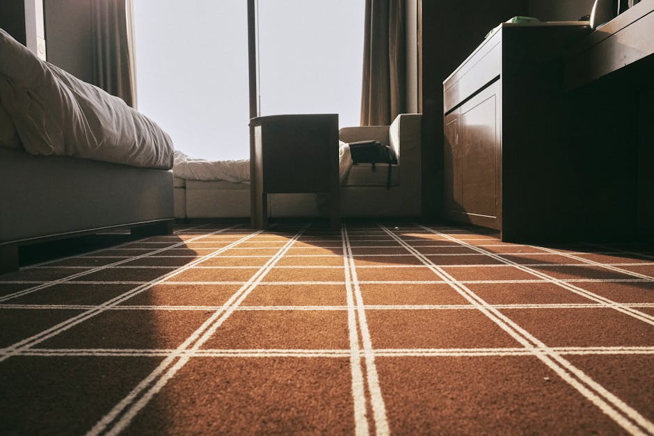 A close-up view of an indoor residential space featuring a brown carpet with a white intersecting line pattern, well-maintained and clean, covering a hardwood floor. To the left, part of a white bed with bedding is visible, and in the background, a tall window with natural light streaming in, partially covered by beige curtains. A dark wooden dresser and a side table are positioned against the wall on the right, with a black telephone placed on top of the side table. The lighting highlights the carpet's smooth texture and the overall tidy appearance of the room, indicative of professional surface cleaning and hygiene maintenance provided by Knightsbridge Carpet Cleaning.