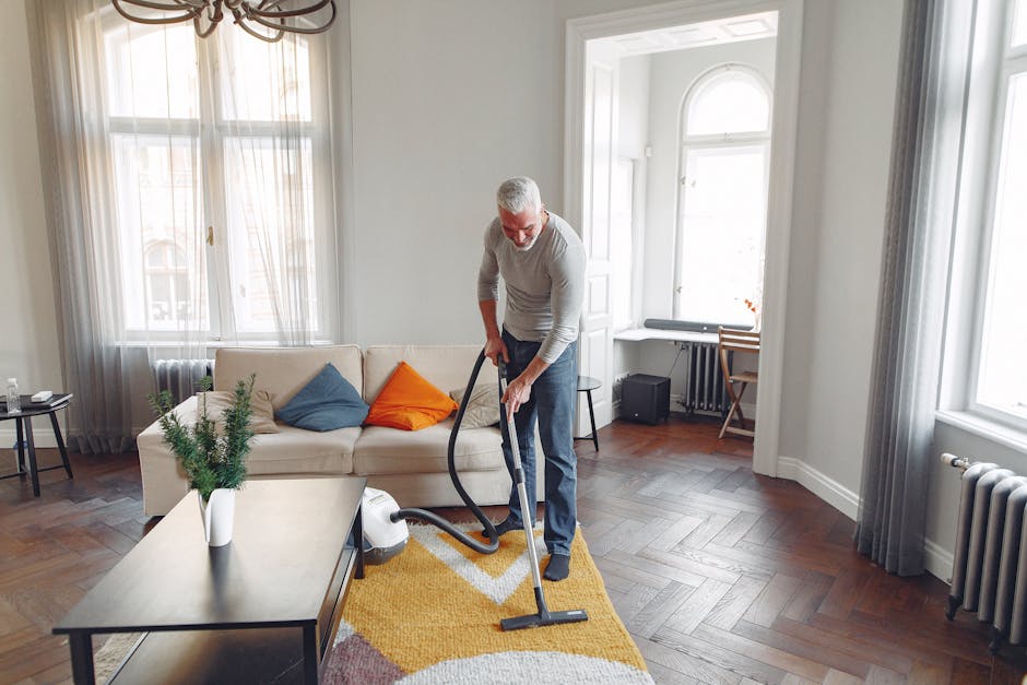 A professional cleaner from Knightsbridge Carpet Cleaning is performing deep surface cleaning in a bright, modern living room. The individual is wearing a full white protective suit, including a hood, gloves, and blue shoe covers, while using a specialized steam or extraction machine connected to a long black hose to thoroughly clean a plush beige rug on the light wood floor. The room features natural light coming through large windows, a wooden shelving unit with decorative items and books, potted plants, and minimal furnishings, emphasizing the clean, organized space. Visible cleaning equipment and the pristine condition of the rug highlight a focus on hygiene and effective carpet maintenance, aligning with residential and commercial cleaning standards.