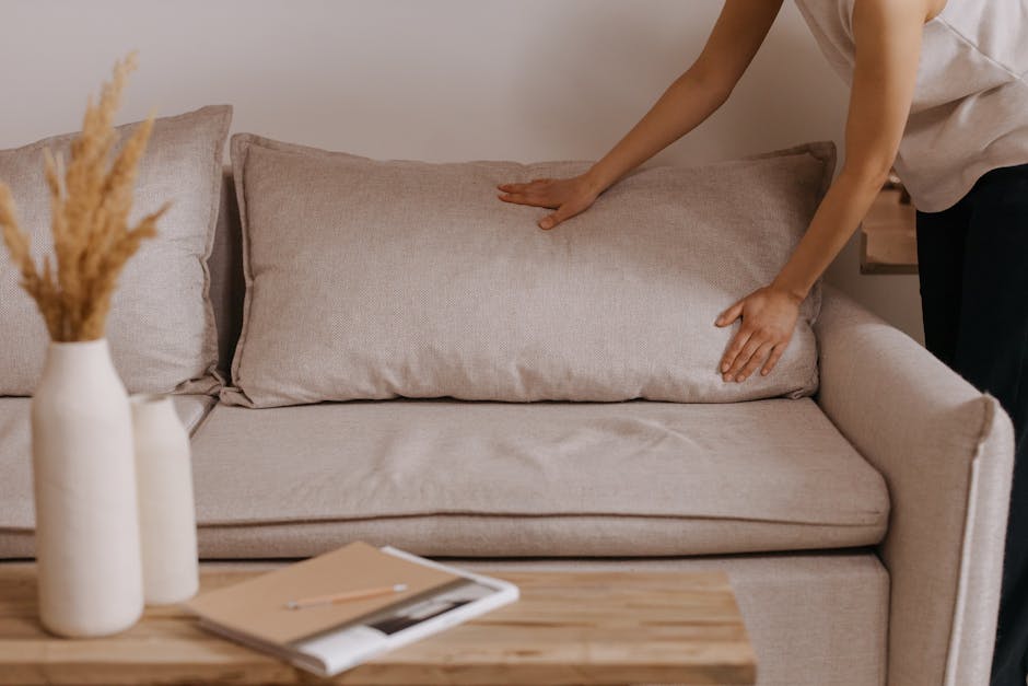 A person adjusting a beige fabric cushion on a light-colored upholstered sofa in a living room. The sofa has a smooth, clean surface with no visible stains or dust. Next to the sofa, two white ceramic vases with dried wheat stems are placed on a wooden coffee table, which also holds an open magazine and a notepad with a pen. The room features soft, natural lighting, and the background shows a plain wall, providing a tidy and well-maintained appearance consistent with professional cleaning standards by Knightsbridge Carpet Cleaning.