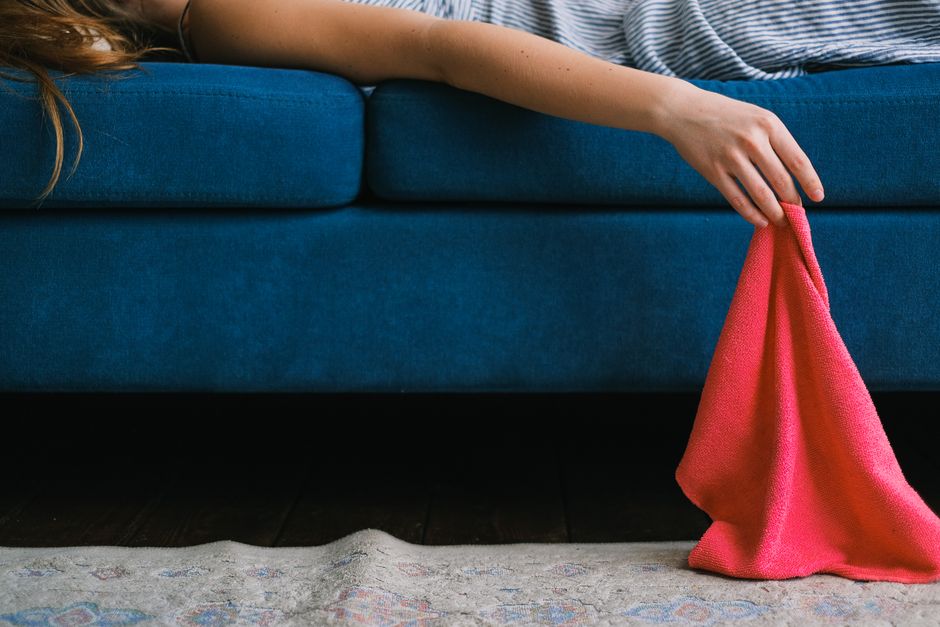 A person adjusting a beige fabric cushion on a light-colored upholstered sofa in a living room. The sofa has a smooth, clean surface with no visible stains or dust. Next to the sofa, two white ceramic vases with dried wheat stems are placed on a wooden coffee table, which also holds an open magazine and a notepad with a pen. The room features soft, natural lighting, and the background shows a plain wall, providing a tidy and well-maintained appearance consistent with professional cleaning standards by Knightsbridge Carpet Cleaning.