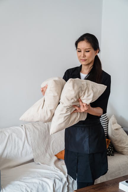 A woman dressed in a black uniform is holding two beige cushions on a white upholstered sofa in a well-lit living room. The sofa has a white fabric cover, with a few other cushions in beige, orange, and black-and-white patterns arranged at one end. The room features white walls and a wooden table or surface partially visible in the foreground. Visible are the cushions' soft fabric material and the clean, tidy appearance of the sofa, indicating recent or ongoing cleaning. The scene emphasizes domestic surface cleaning and maintenance, consistent with the services offered by Knightsbridge Carpet Cleaning, particularly in relation to upholstery cleaning in Knightsbridge flats.