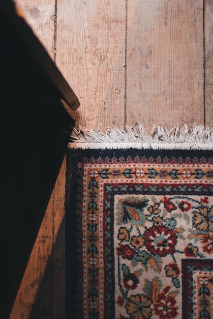 Close-up of a patterned area rug with intricate floral and geometric designs in red, blue, beige, and green, situated partially under a dark piece of furniture on a hardwood floor. The wooden flooring features visible grain patterns and a light, natural finish, with some minor wear and color variation. The rug's fringe is seen along the edge, and the lighting is soft, highlighting the texture and cleanliness of the surfaces. This setup exemplifies interior surface cleaning and maintenance, consistent with Knightsbridge Carpet Cleaning's domestic cleaning services highlighted in the SW1X rug cleaning guide for Knightsbridge, London.