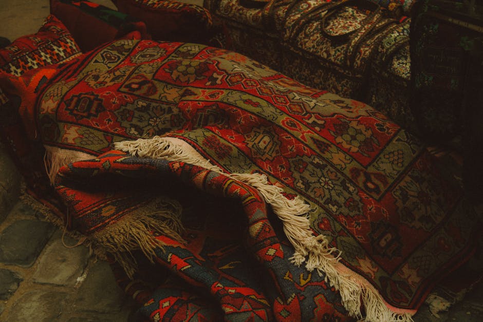 Close-up of a patterned area rug with intricate floral and geometric designs in red, blue, beige, and green, situated partially under a dark piece of furniture on a hardwood floor. The wooden flooring features visible grain patterns and a light, natural finish, with some minor wear and color variation. The rug's fringe is seen along the edge, and the lighting is soft, highlighting the texture and cleanliness of the surfaces. This setup exemplifies interior surface cleaning and maintenance, consistent with Knightsbridge Carpet Cleaning's domestic cleaning services highlighted in the SW1X rug cleaning guide for Knightsbridge, London.
